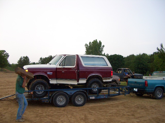 Maroon and white Ford Bronco on a trailer, being loaded by a person carrying wood. Other trucks are visible in the background