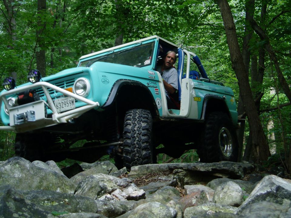 Teal Ford Bronco with white top and roll cage, off-roading over rocks