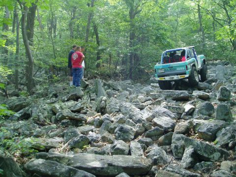 1969 Ford Bronco - A teal Ford Bronco tackles a challenging rocky terrain in a wooded area.
