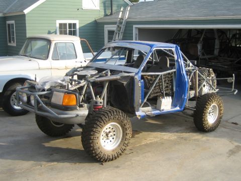 1990 Ford Bronco - blue tube-chassis off-road race truck under construction sits in a driveway next to a vintage white pickup and garage.