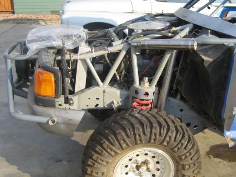 1990 Ford Bronco - partially stripped vehicle front clip showing the bare frame, tubular bumper, an amber turn signal, a red coil spring and a knobby off-road…