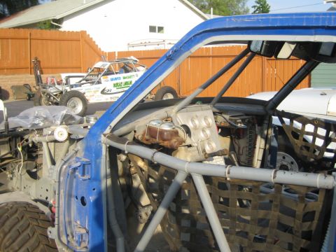 1990 Ford Bronco - View through the roll-cage interior of a blue-framed race truck, with a sponsor-covered off-road racer parked in the background by a wooden…