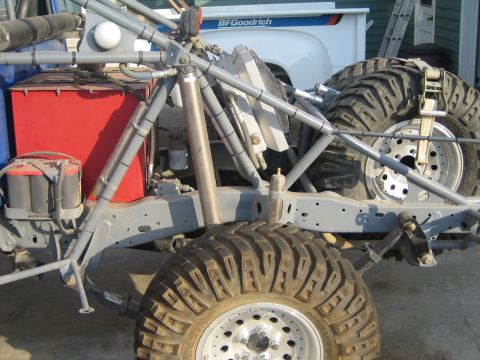 1990 Ford Bronco - stripped vehicle frame showing a red fuel cell, suspension links and large knobby tires, with a white pickup bearing a BFGoodrich sticker b…