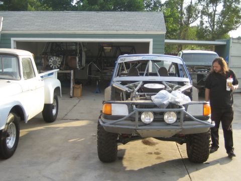 1990 Ford Bronco - white tube-frame off-road race truck numbered 742 sits in a driveway next to a classic white pickup, with a person holding a drink standing…
