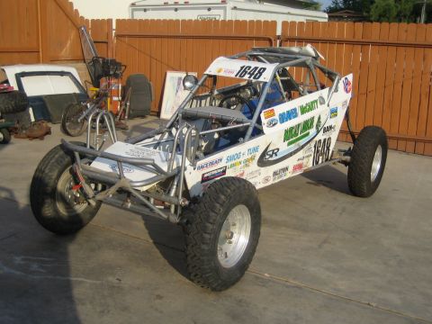 1990 Ford Bronco - white tube-frame off-road buggy covered in sponsor decals and marked 1648 sits on knobby tires against a wooden fence.