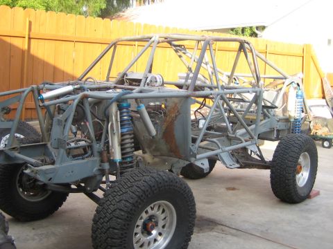 1990 Ford Bronco - bare gray tube-frame off-road buggy chassis with blue coilover shocks and knobby tires sits in a yard against a wooden fence.