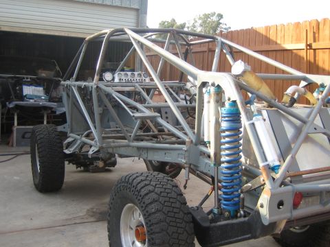 1990 Ford Bronco - Side profile of an unpainted tube-frame off-road buggy showing a dashboard cluster, a tall blue coilover shock and knobby tires.