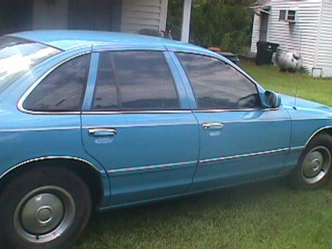 1994 Ford Crown Victoria - The side profile of a blue 1994 Ford Crown Victoria sedan is visible against a backdrop of a house and lawn.
