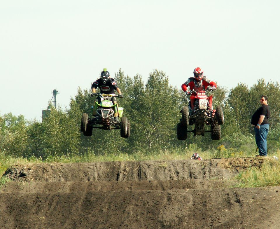 Two ATVs, one green and one red, airborne over a dirt motocross jump with trees behind