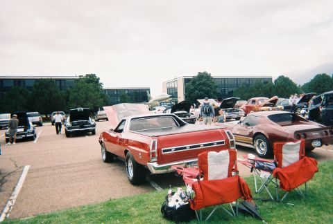 1972 Ford Ranchero - A red Ford Ranchero pickup truck is displayed at an outdoor car show with other vehicles and buildings in the background.