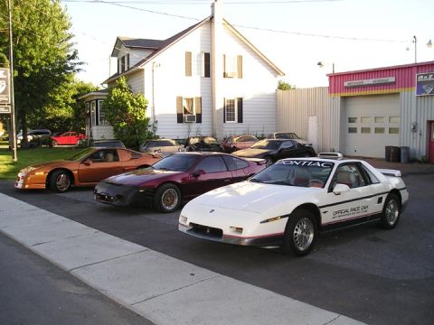 1984 Pontiac Fiero - Several Pontiac Fiero models are lined up in front of a building, with the closest one being a white 'Official Race Car' edition.