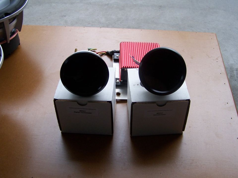 Two black gauge pods and a red electronics module on cardboard boxes for tach and speedo