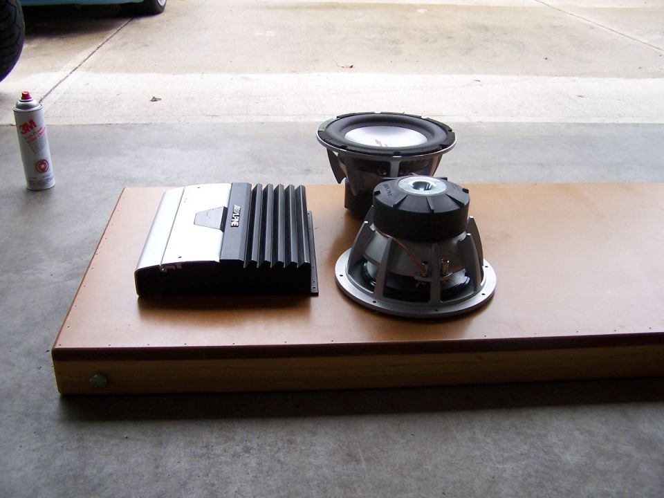 Car amplifier and two subwoofers laid out on a wood board in a garage driveway