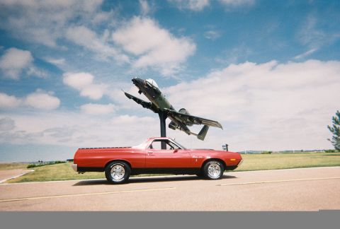 1972 Ford Ranchero - A red pickup truck is parked on a road with a jet fighter plane displayed overhead against a cloudy blue sky.
