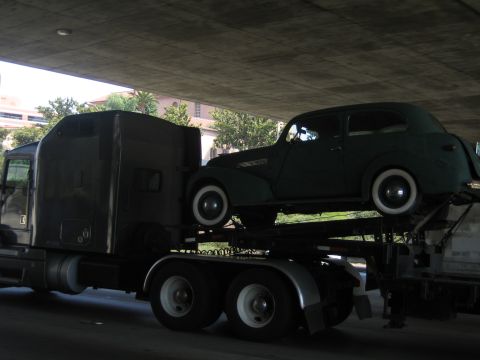 1990 Ford Bronco - dark green vintage two-door sedan with whitewall tires rides atop a car-hauler semi-truck passing beneath a concrete overpass.