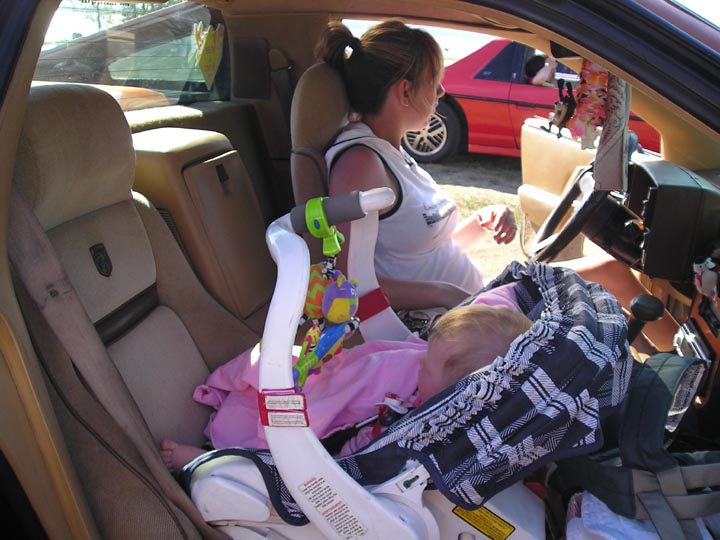 1984 Pontiac Fiero interior with baby in car seat and woman in driver's seat. Red Fiero visible outside