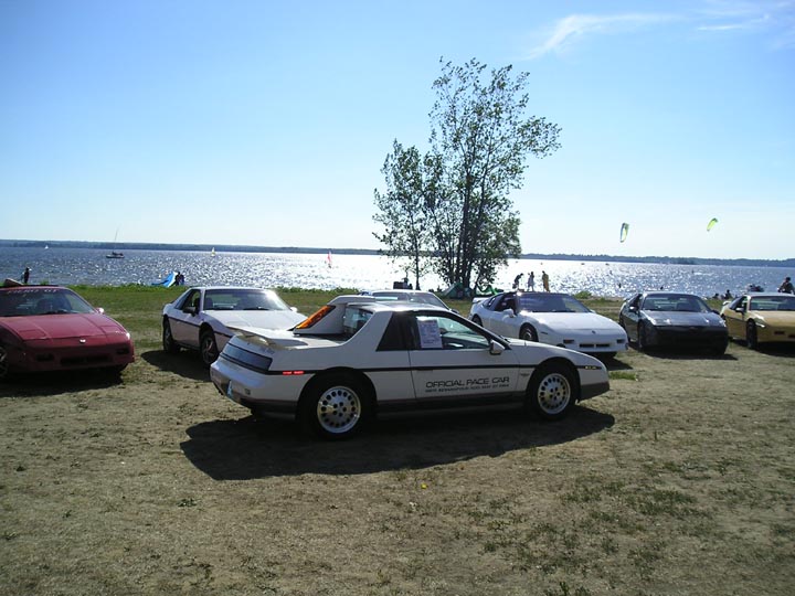 Several 1984 Pontiac Fieros parked near a lake on a sunny day. One white Fiero has 'OFFICIAL PACE CAR' text on its door