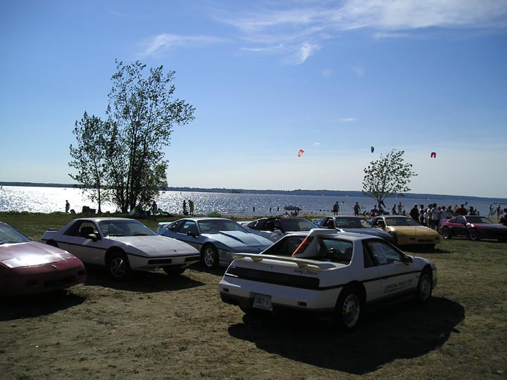 Several Pontiac Fieros parked near a body of water on a sunny day. One Fiero has 'Indy Fiero' text on its rear quarter panel
