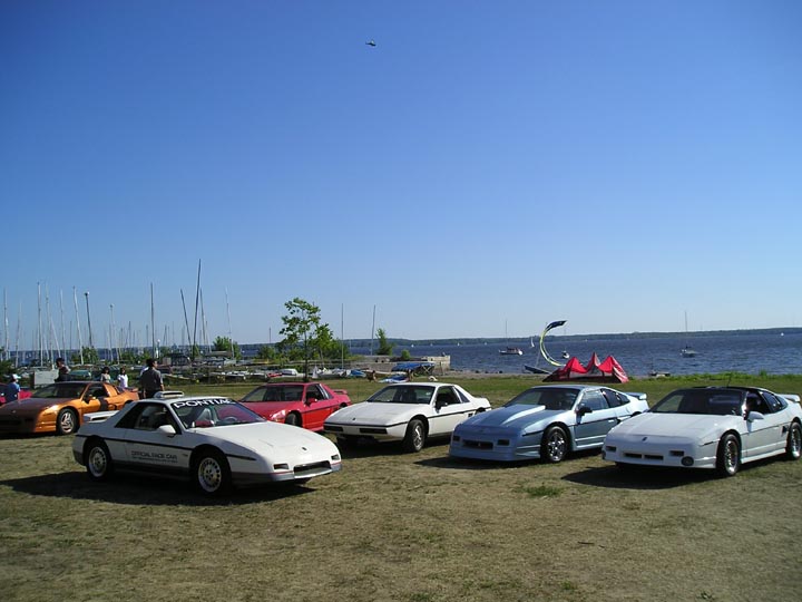 White 1984 Pontiac Fiero with 'Pontiac Pace Car' text on windshield, parked on grass near a body of water with sailboats