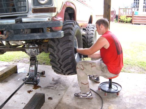 1978 Ford Bronco - A man is working on the front wheel of a heavily modified truck that is supported by a jack.