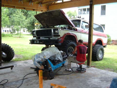 1978 Ford Bronco - A man works on the engine of a lifted 1978 Ford Bronco parked under a wooden structure.