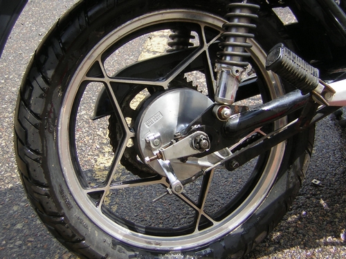 Close-up of motorcycle rear alloy wheel with drum brake hub, brake rod, shock absorber, and footpeg on wet pavement