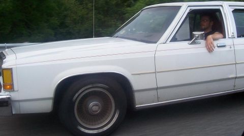 1985 Ford LTD Crown Victoria - A white 1985 Ford Crown Victoria LTD sedan is seen from the side, with a driver visible through the window.