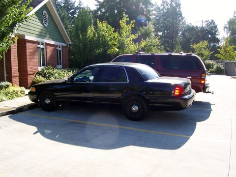 2003 Ford Crown Victoria - A black sedan is parked in a lot next to a red SUV, with a building and trees visible.