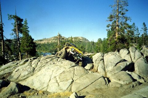 1967 Ford Bronco - A yellow Jeep Wrangler navigates a rocky trail near a lake, with mountains and pine trees in the distance.