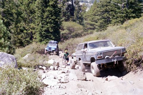 1967 Ford Bronco - Two off-road vehicles navigate a challenging rocky path through a wooded area.