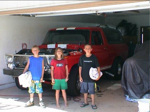 1979 Ford Bronco - Three children pose in front of a red Ford Bronco in a garage, with the hood up and another car covered nearby.