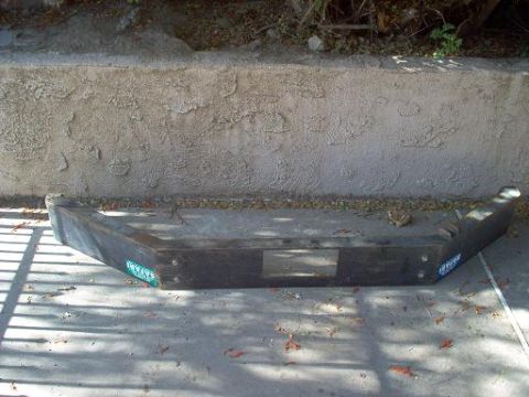 1988 Ford Bronco - A bare steel front bumper laid face-up on a concrete driveway against a stucco retaining wall, its dark weathered finish showing a flat lic…
