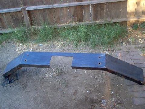 1988 Ford Bronco - A fabricated flat steel bumper shell sitting on bare dirt in front of a wooden fence, with a large rectangular cutout in the center face fo…