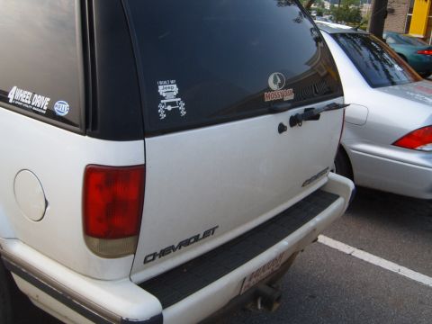 1988 Ford Bronco - A white Chevrolet SUV is parked next to a silver sedan, displaying several decals on its rear window.