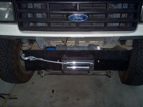 1988 Ford Bronco - Head-on view of a white Ford truck's front end in a garage, showing a winch mounted behind a black fabricated bumper/mounting plate below t…