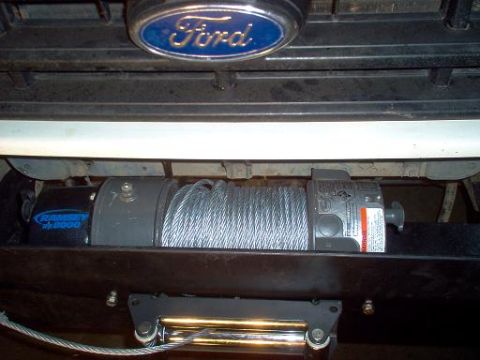 1988 Ford Bronco - A Ramsey electric winch with steel cable spooled on its drum is mounted behind the front bumper of a Ford truck, the blue Ford oval badge o…