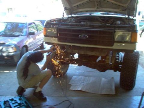 1988 Ford Bronco - A person uses an angle grinder on the front suspension of a lifted Ford truck, with another vehicle in the background.
