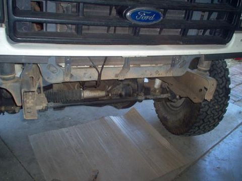 1988 Ford Bronco - Low front view of a white Ford truck with the bumper off, showing the bare frame rails, front crossmember, radius arm brackets and steering…