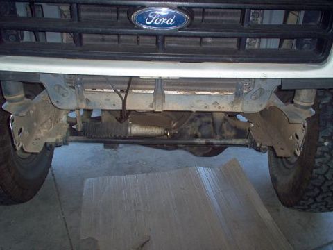 1988 Ford Bronco - Straight-on low view of a white Ford truck with the front bumper removed, exposing both frame horns, the front crossmember, radius arm brac…