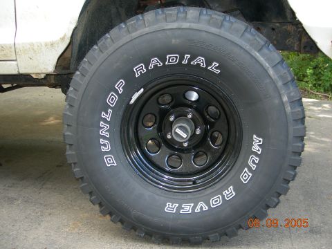 1984 Ford Bronco II - A close-up view of a Dunlop Mud Rover radial tire mounted on a black wheel, with a portion of a white vehicle in the background.