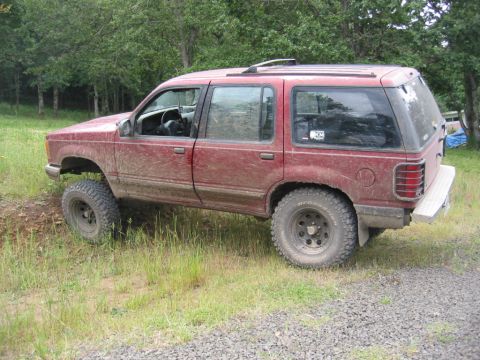 1991 Ford Explorer - This red SUV is shown off-road with its tires on uneven ground and grass.