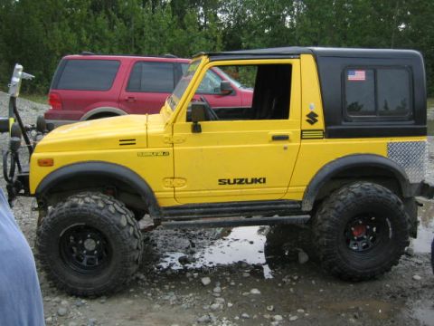 1987 Suzuki Samurai - A yellow 1987 Suzuki Samurai is parked next to a red SUV on a dirt lot.