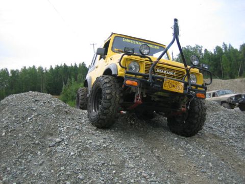 1987 Suzuki Samurai - A yellow 1987 Suzuki Samurai is shown off-road on a gravel incline with other vehicles in the background.