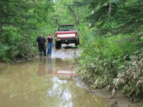 1987 Suzuki Samurai - A red pickup truck navigates a muddy path through a dense forest, with two individuals observing its progress.
