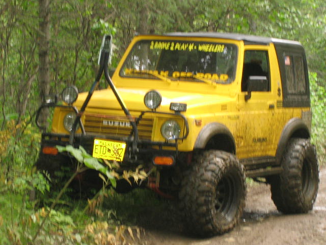 Yellow lifted Suzuki Samurai on mud tires with brush guard on a forest dirt trail