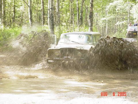 1987 Suzuki Samurai - Two off-road vehicles navigate a muddy trail in a wooded area, creating large splashes.