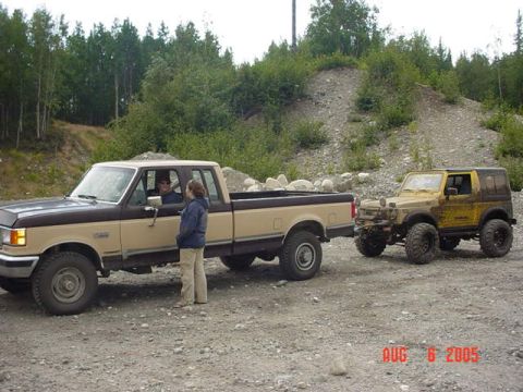 1987 Suzuki Samurai - Two off-road vehicles are parked on a gravel road with trees and a hill in the background.