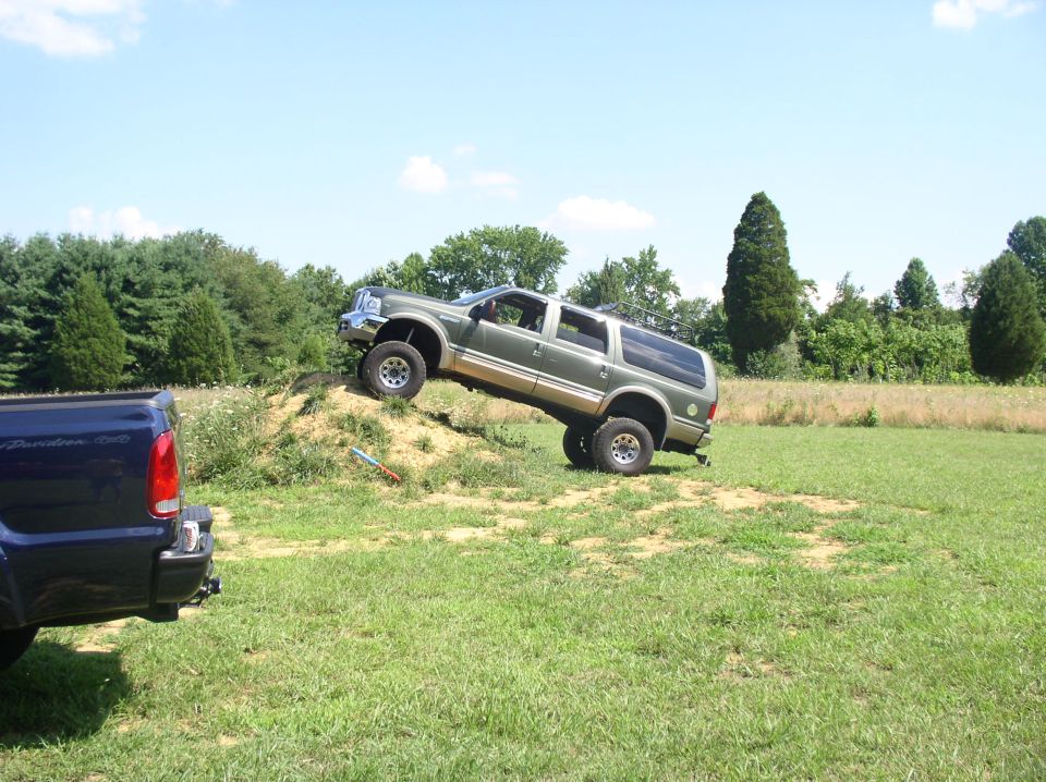 Lifted tan Ford Excursion SUV climbing dirt mound in grass field, blue pickup bed in foreground