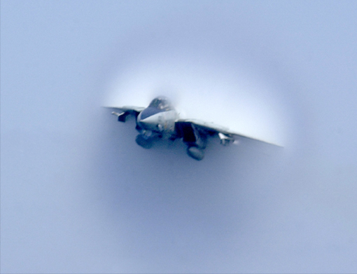 Fighter jet with vapor cone forming around it against a blue sky