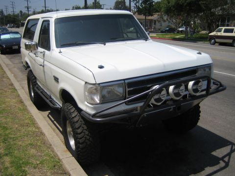 1990 Ford Bronco - white lifted Ford Bronco full-size SUV with a tubular brush bumper, round driving lights and off-road tires parked along a residential curb.